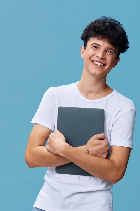 smiling man holding laptop, casual white t-shirt, curly hair, blue background, young adult with joyful expression, modern technology lifestyle concept.