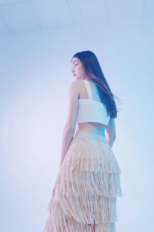 Young woman in a stylish white top and fringed skirt poses against a soft blue background. The image highlights beauty, fashion, and elegance in a modern studio setting.