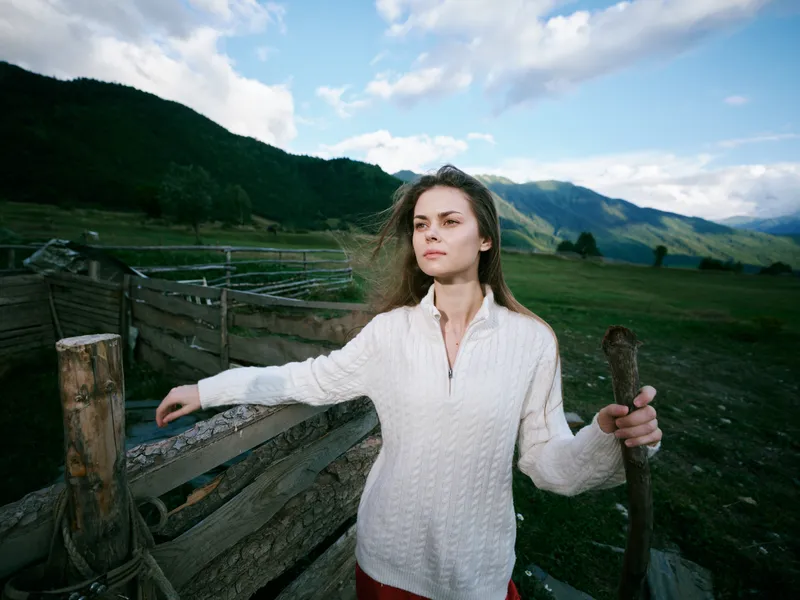Woman leaning on a wooden fence in a rural countryside, wearing a white sweater, gazing toward distant hills under a blue sky, capturing calm, rustic life and nature