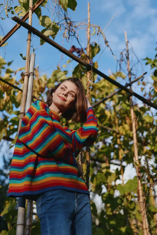 Woman in rainbow striped sweater portrait in garden, smiling and posing among trellis vines, casual autumn fashion with natural light, relaxed outdoor lifestyle and seasonal foliage.