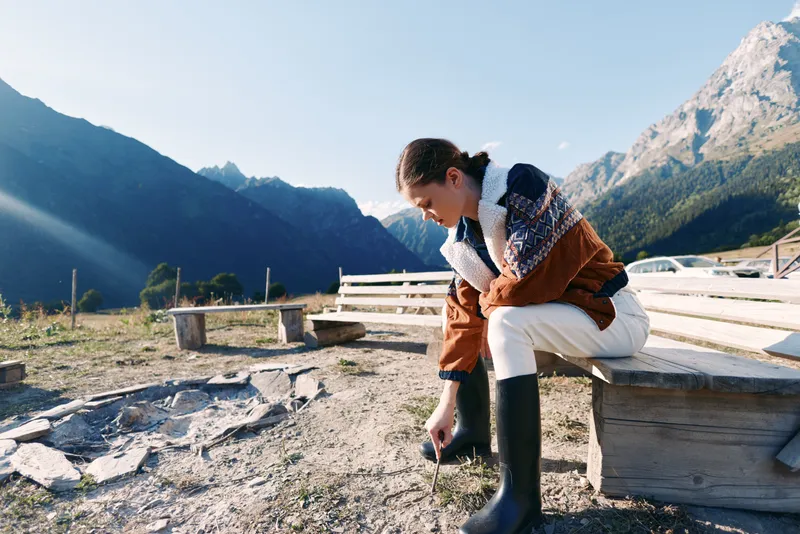 Woman in warm sweater sits on wooden bench outdoors by mountain lake, tying black rubber boots, enjoying sunlight and alpine scenery during cool hiking or countryside break.