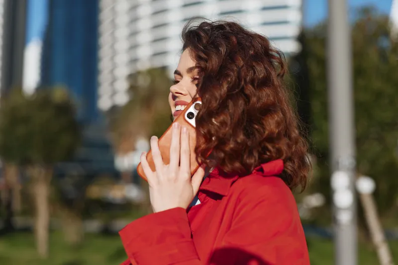 Young woman with curly hair smiles happily while talking on a smartphone outdoors in a bright red coat during a sunny day in an urban park.
