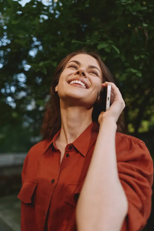 Woman on outdoor call, smiling, wearing a red shirt, enjoying a sunny day, nature background, relaxed lifestyle moment, candid portrait, cheerful mood during conversation
