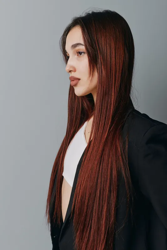 Young woman with long straight red hair wearing black jacket and white top standing in profile against gray background for beauty fashion portrait.