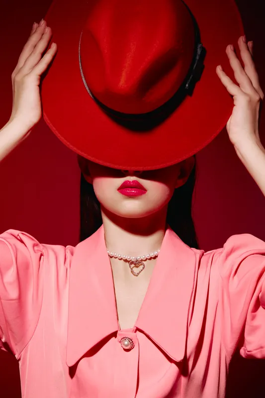 Fashionable woman with bright red hat covering eyes wears pink outfit with pearl heart necklace against red background. Bold lip color and stylish composition. Beauty and fashion concept.