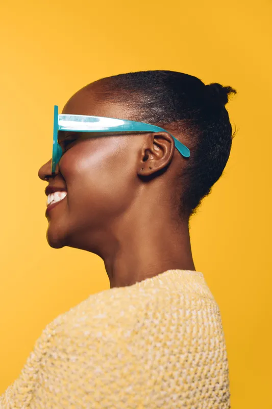 Profile of a smiling black woman with dark skin wearing blue sunglasses and a yellow sweater. Bright yellow background creates a vibrant, cheerful mood and modern fashion concept.