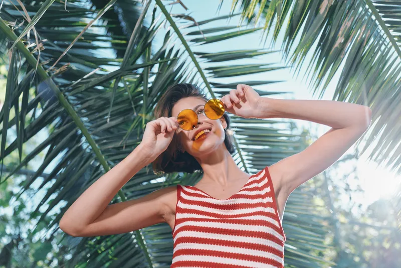 A cheerful woman wearing a striped tank top and sunglasses enjoys the sunny outdoors among lush palm leaves, radiating carefree energy and vibrant summer vibes for stock photography.