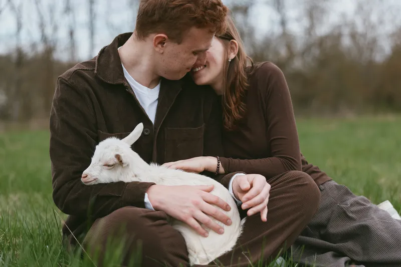 A couple sits in a grassy field outdoors, warmly embracing a small goat kid resting on their laps, with tender contact and bright smiles conveying simple lifestyle joys and animal companionship.