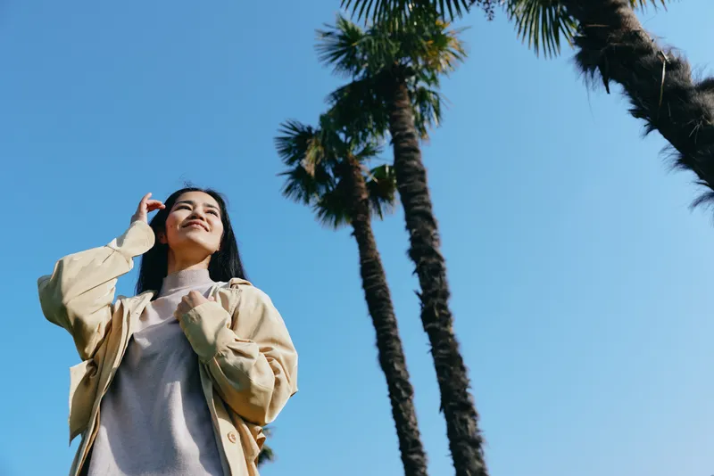 Sunlit scene featuring a woman looking up toward a clear blue sky beside tall palm trees, wearing a light jacket, conveying freedom, outdoor travel, and carefree mood.