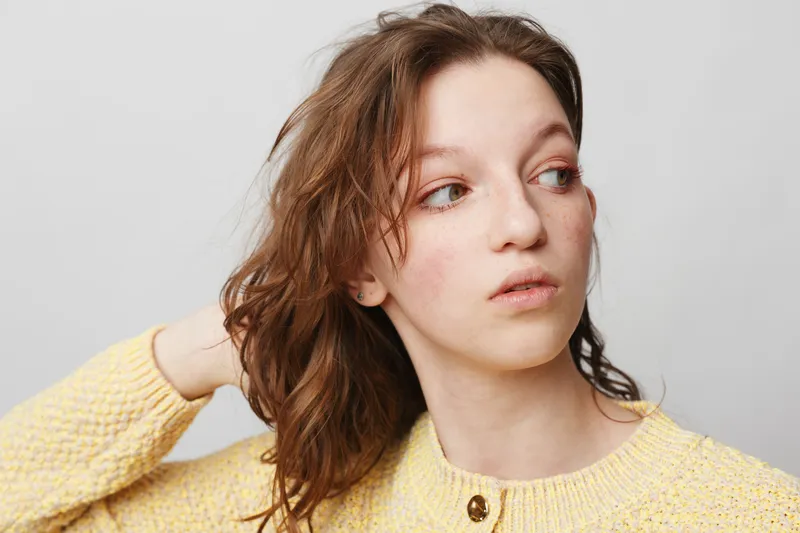 Young woman with wet brown hair wearing yellow sweater looks away thoughtfully on light background. Concept of natural beauty, casual style, and introspection in portrait photography.