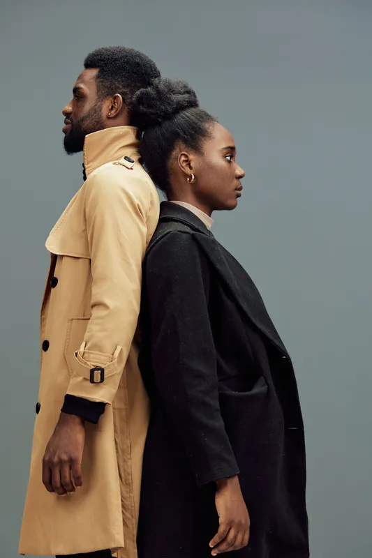 Stylish couple in contemporary fashion standing back to back, showcasing modern clothing and confident expressions against a muted gray background