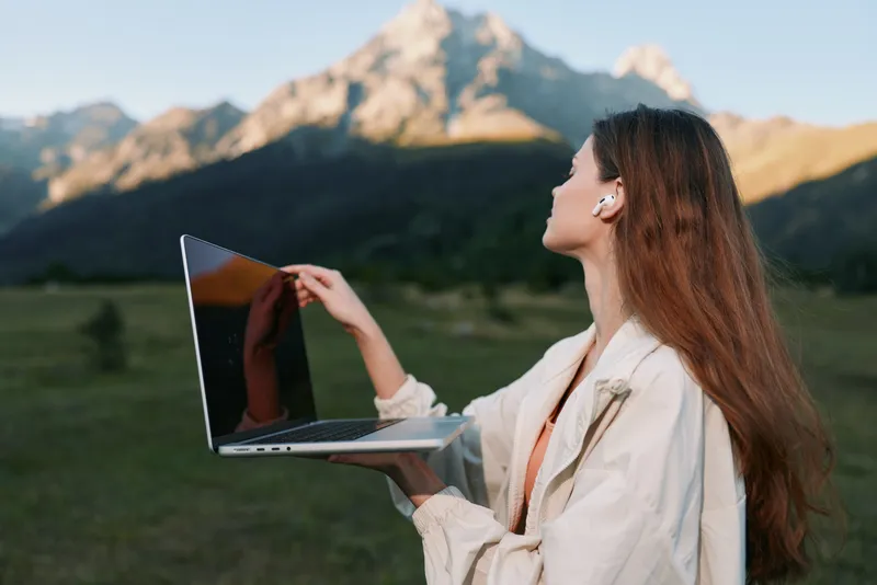 A professional woman stands outdoors in a scenic mountain landscape, holding a laptop and looking at the screen with a focused, engaging expression.