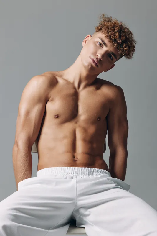 Young man with curly hair posing shirtless in white pants against gray background, showing muscular torso and relaxed confident expression in studio portrait.