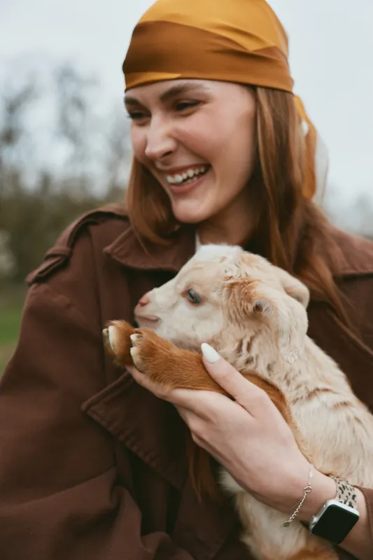 Smiling woman wearing a headscarf holds a playful goat kid outdoors, showcasing a lifestyle moment of bonding, care, and rural charm, with warm, natural light.