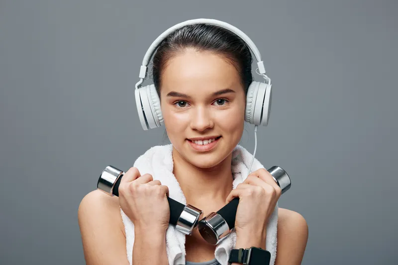 Fit woman lifts dumbbells with a towel around her neck and headphones, performing strength training in a clean gym, showcasing focus, balance and athleticism during a workout.