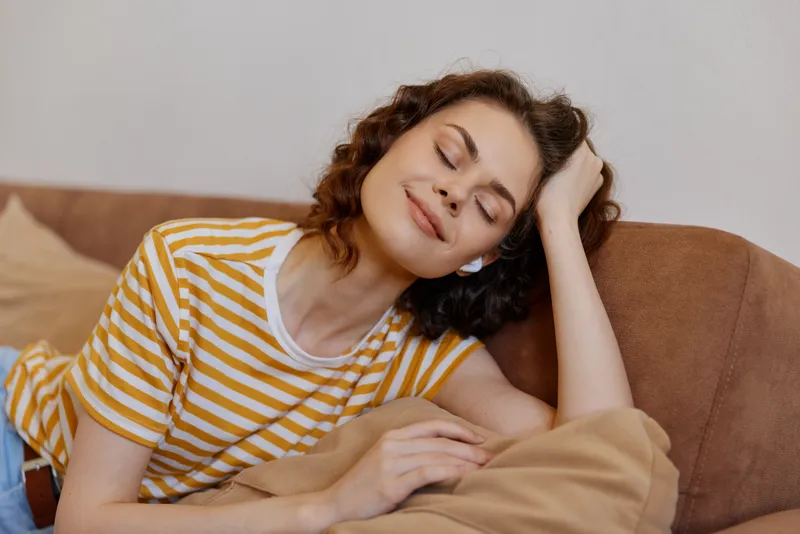 Young woman relaxes on a sofa in a casual striped tee, with a calm smile, enjoying a peaceful moment of comfort at home.