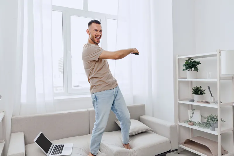 Joyful man dancing on sofa in cozy living room, embodying authentic imperfection and embracing mental well-being while enjoying a digital detox experience.
