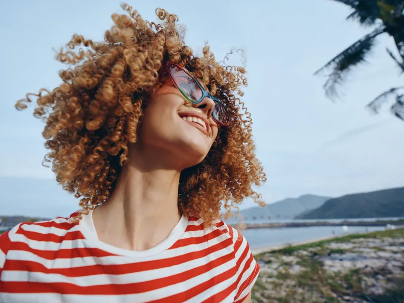 Joyful portrait of a smiling woman with voluminous curly hair, striped shirt, and sunglasses, captured outdoors near a sunny beach with palm trees and breeze.