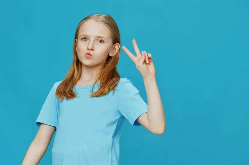 Cheerful girl with long hair making a peace sign against a bright blue background, wearing a light blue t shirt and expressing playful emotions