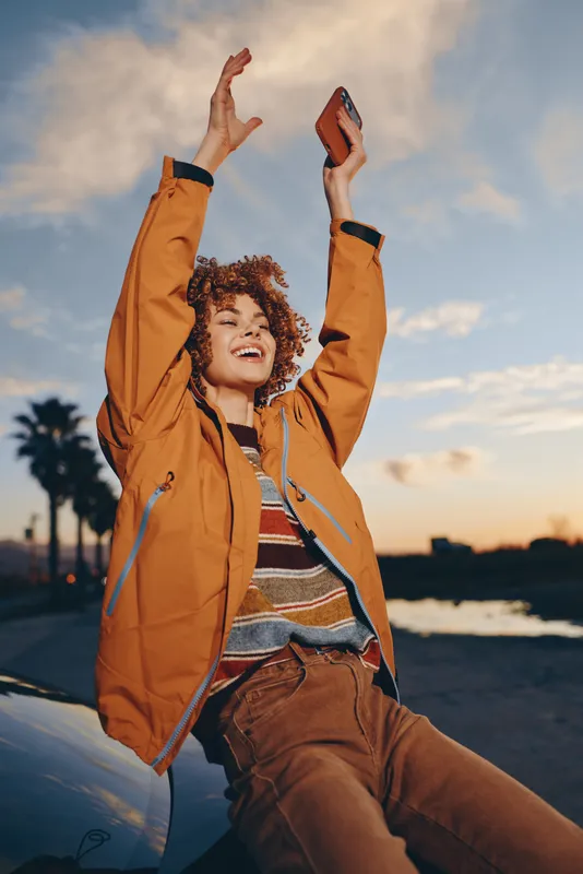 Woman lifestyle portrait at sunset beach, joyful curly haired woman in orange jacket holding smartphone overhead, laughing outdoors with arms up, warm evening light and ocean horizon