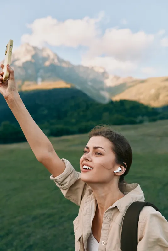 Young woman lifestyle outdoors, smiling and holding phone up, wireless earbuds, casual clothing, mountain landscape background, sunny day, natural light