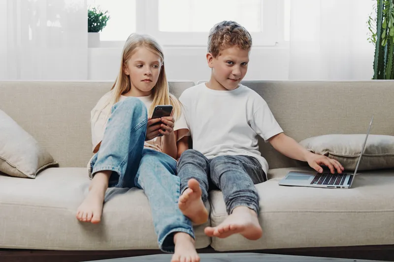 Two children sitting on a beige sofa using digital devices, a girl holding a smartphone and a boy typing on a laptop. Natural light brightens the modern living room scene.