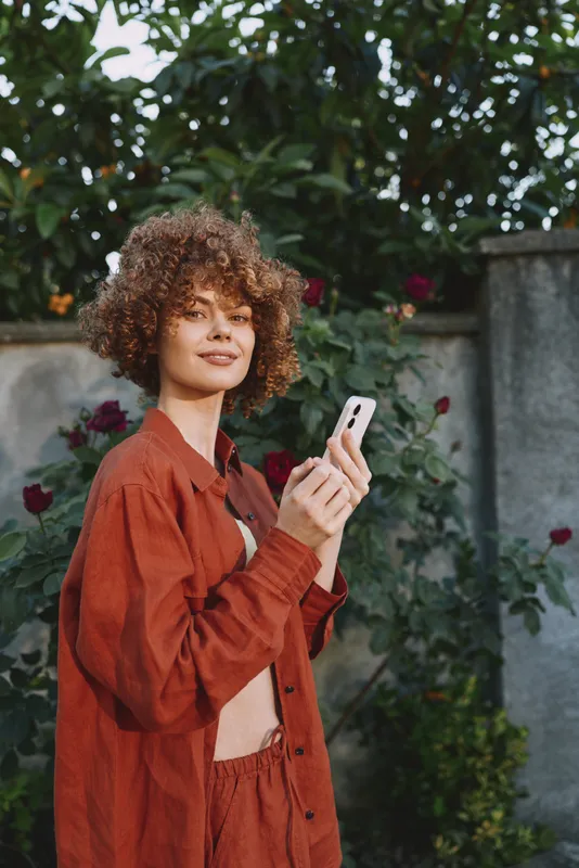 Young woman with curly hair holds a smartphone in a garden, wearing a rust jacket and a relaxed, confident smile