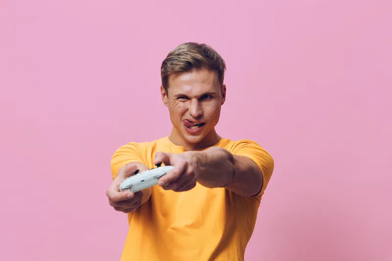 Smiling man with short brown hair wearing yellow T-shirt holding game controller, looking excited and focused, isolated on pink background, studio portrait, entertainment lifestyle concept, gaming moo