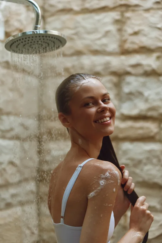 smiling woman showering outdoors under rainfall, wet hair, natural stone wall background, relaxed mood, morning light, casual white top