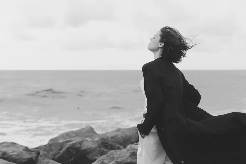 Windblown coat on a lone woman by the ocean, standing on rocks with the coast behind, mood of solitude and freedom in monochrome scenery for dramatic fashion