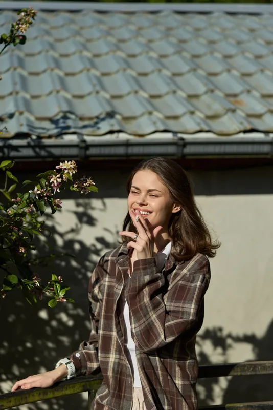 Young woman in a plaid shirt smiles softly in daylight, standing beside a wall with flowering branches and a tiled roof, capturing a candid, relaxed lifestyle moment