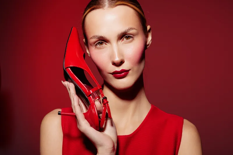 Woman in red dress holds red high heel shoe near face against red background. Fashion concept with bold makeup and elegant style, highlighting color coordination and glamour.