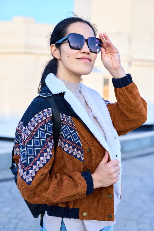 Female in patterned jacket and backpack adjusts sunglasses while posing outdoors, showcasing streetwear fashion, confident style and a bright smile in an urban setting.