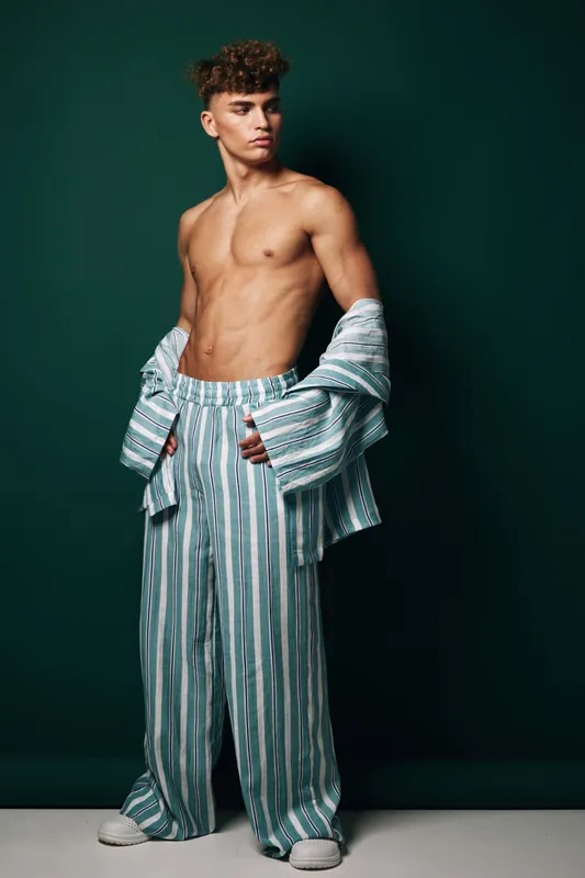 Young man with curly hair posing shirtless wearing striped wide pants and open shirt against dark green background in studio for fashion and style concept.