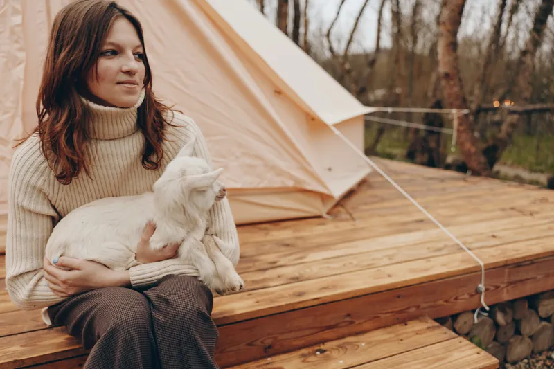 Young woman in a beige turtleneck sweater sits on wooden steps holding a white baby goat near a tent in an autumn outdoor setting with calm expression.