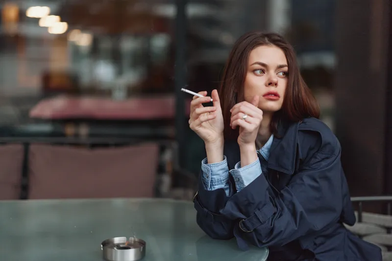 Brunette woman in a denim jacket sits at an outdoor cafe, smoking a cigarette, looking thoughtful as warm evening light and city glow surround her.