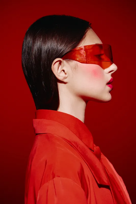 Profile portrait of young woman wearing red fashion coat and translucent red sunglass, modern beauty style with glossy lips and vibrant blush on red background.
