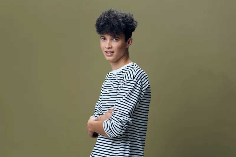 young man in striped shirt posing confidently against olive green background with curly hair and casual style, showcasing modern youth fashion and relaxed attitude