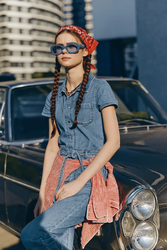 stylish young woman with sunglasses and braided hair leans on classic black car outdoors in urban setting, wearing denim and red headscarf in sunny daylight