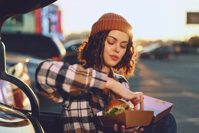 Woman with burger in takeaway box by car trunk at sunset, beanie and plaid jacket create a cozy outdoor scene with authenticity, candid lifestyle, golden hour glow, mindful living, emotional