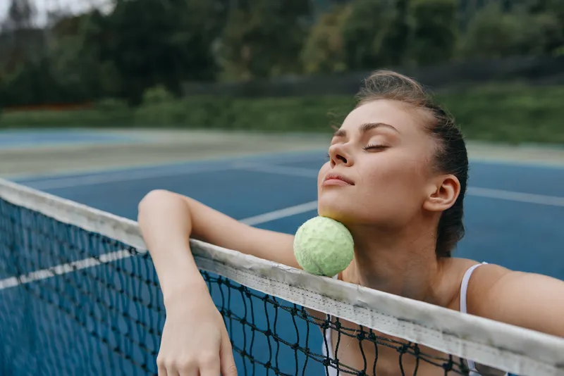 Young athletic woman enjoying a moment of relaxation with a tennis ball on her shoulder, set on a serene blue tennis court surrounded by greenery