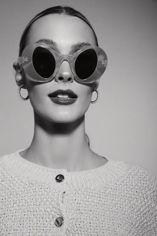 Stylish light skinned woman wearing large round sunglasses and hoop earrings poses confidently in cozy knitted sweater.