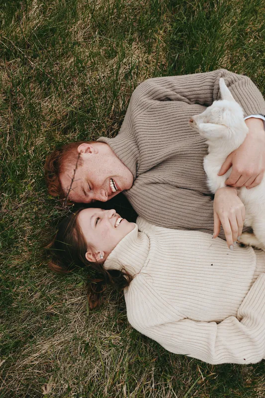A couple relaxes on grass in cozy sweaters, smiling as they cradle a white goat kid. A warm, natural lifestyle scene featuring animal companionship and gentle affection.