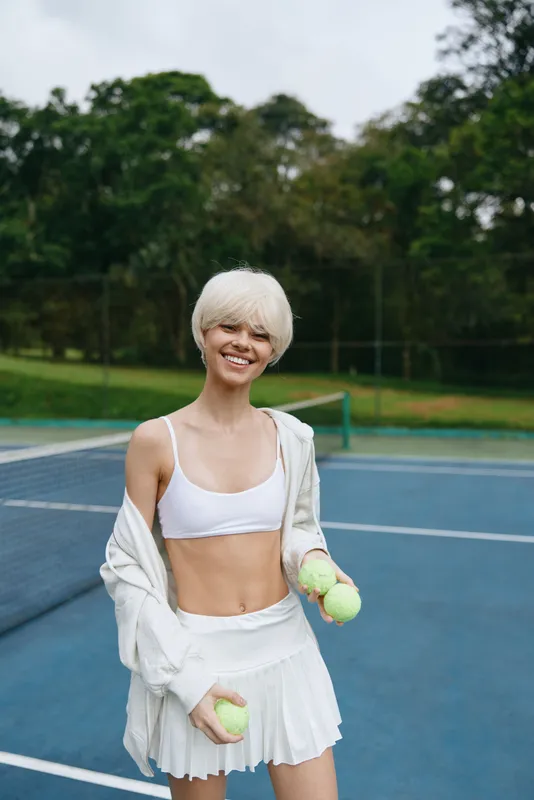 Young athletic woman with short blonde hair smiling while holding tennis balls on a sunny court, showcasing an active lifestyle and sportswear fashion