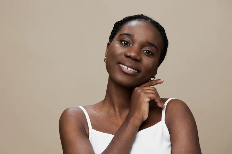 A confident young Black woman smiling with a hand under her chin, wearing a white top, against a neutral background, showcasing natural beauty and elegance