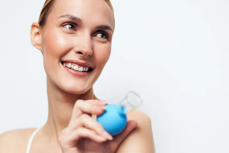 A smiling young woman with clear skin holds a blue facial cleansing brush near her face. The bright background highlights skincare and beauty routine concepts.