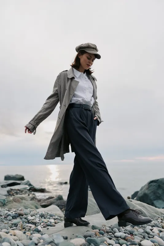 Young woman wearing stylish coat and cap walks confidently on rocky beach near calm sea under cloudy sky. Concept of fashion, autumn, and outdoor lifestyle with natural light.