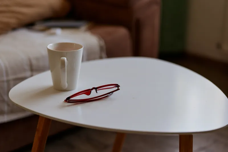 Cozy home living scene with a white mug and red glasses resting on a round table, soft lighting and a blurred sofa backdrop, conveying morning routine and quiet relaxation and calm mood.