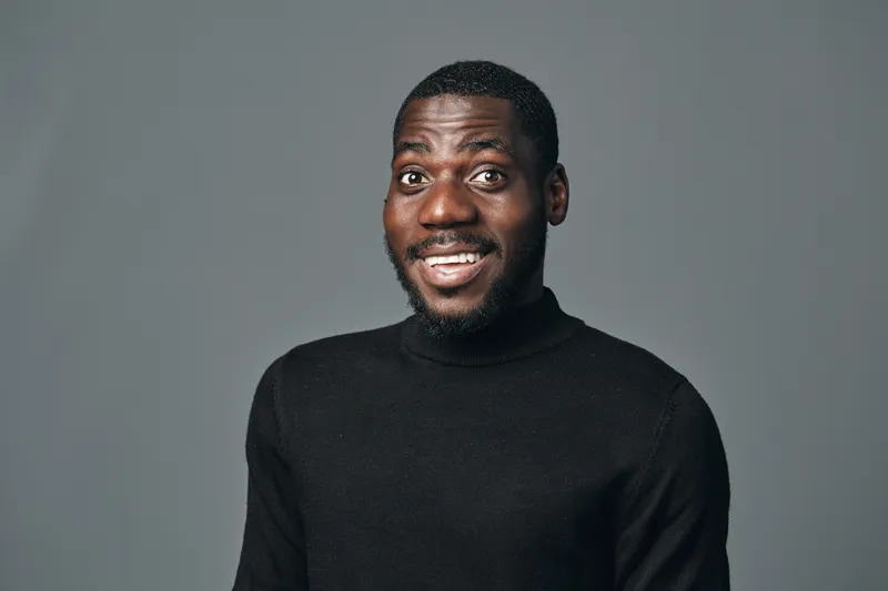 African man with dark skin wearing black turtleneck sweater posing with surprised expression in creative conceptual visual style on gray background. Studio portrait of young male model.