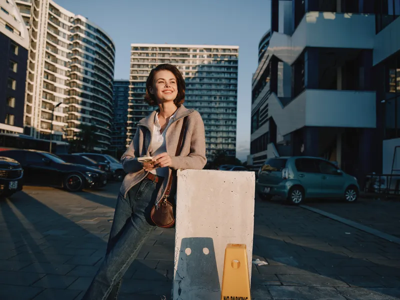 A cheerful woman stands outside in an urban setting, smiling while texting on her smartphone. Natural daylight highlights her confident pose as modern architecture and parked cars create a vibrant